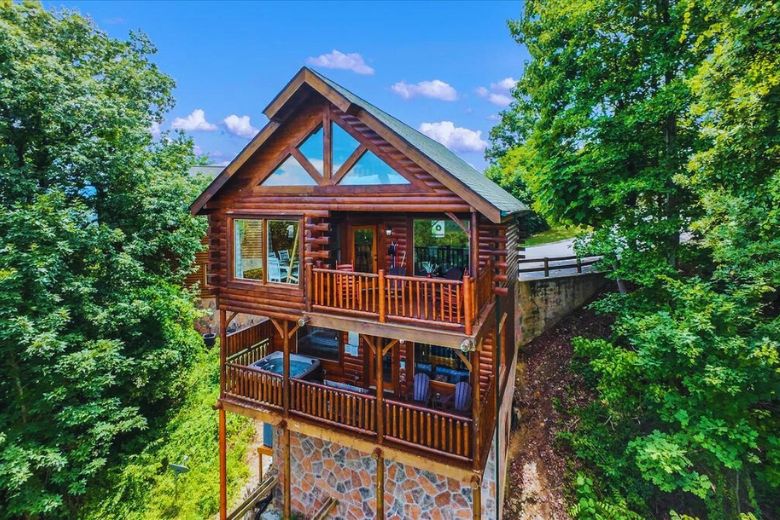 Three-story log cabin with large windows and multiple balconies nestled in a wooded hillside, featuring stonework on the lower level and surrounded by lush green trees under a bright blue sky.